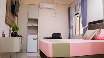 A hotel room with a bed, a desk, a chair, a small refrigerator, a TV mounted on the wall, and a window with curtains.
