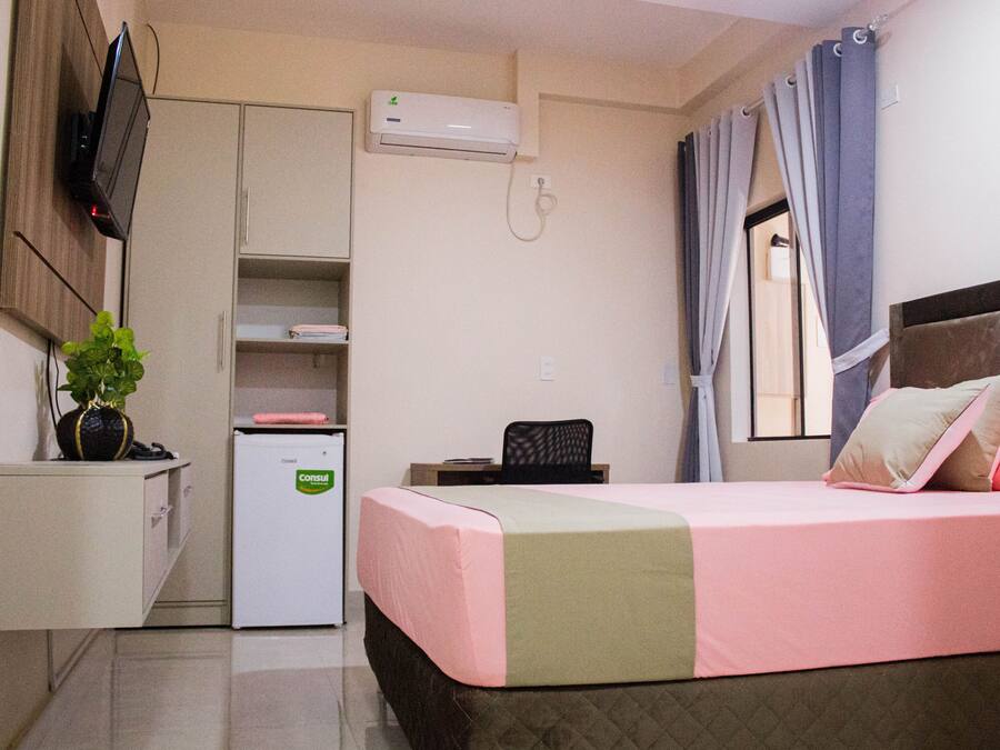 A hotel room with a bed, a desk, a chair, a small refrigerator, a TV mounted on the wall, and a window with curtains.