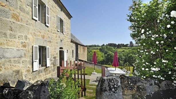 A stone building with shutters, a patio with chairs and umbrellas, and a garden with white flowers. - Gîtes de France 6 people in Le Teil (ALBARET LE COMTAL)