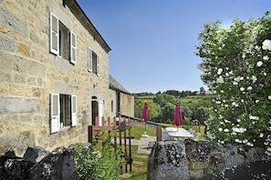 Un bâtiment en pierre avec des volets, une terrasse avec des chaises et des parasols, et un jardin avec des fleurs blanches.