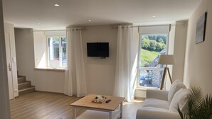 En moderne stue med trægulv, hvid sofa og sofabord. Et fjernsyn er monteret på væggen, og der er vinduer med gardiner.