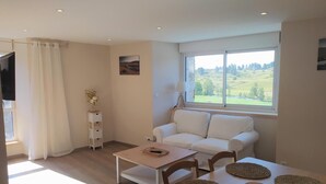 Un salon moderne avec un canapé blanc, une table à manger avec des sets de table, un petit meuble blanc et une fenêtre donnant sur un paysage verdoyant.