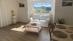 Un salon lumineux avec un canapé blanc, une table basse et une table à manger agrémentée de sets de table. Une fenêtre offre une vue sur un paysage verdoyant.