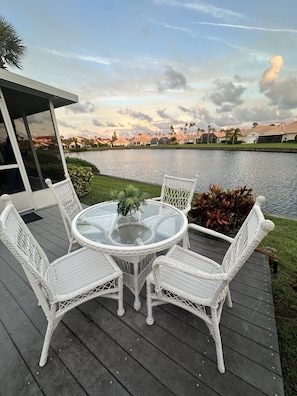 A deck with a round glass table and white wicker chairs, overlooking a body of water and a residential area.