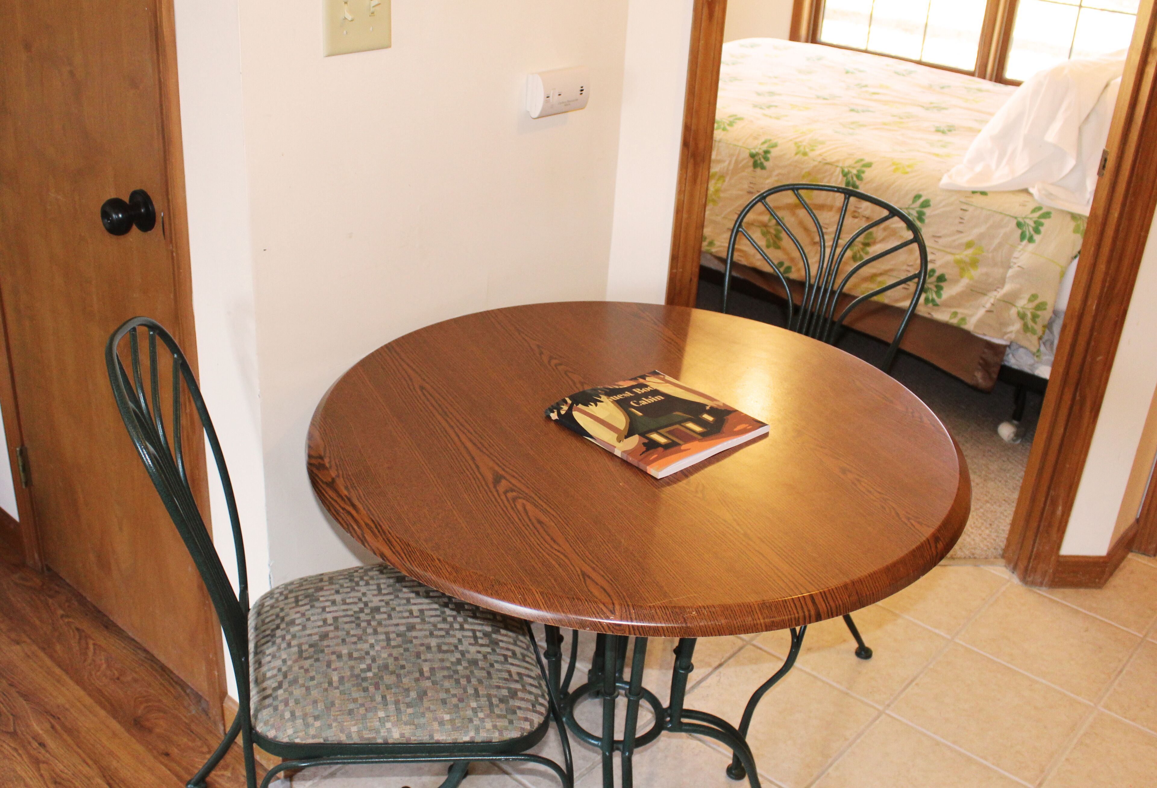 A small round wooden table with a chair, a book on the table, and a bed in the background.