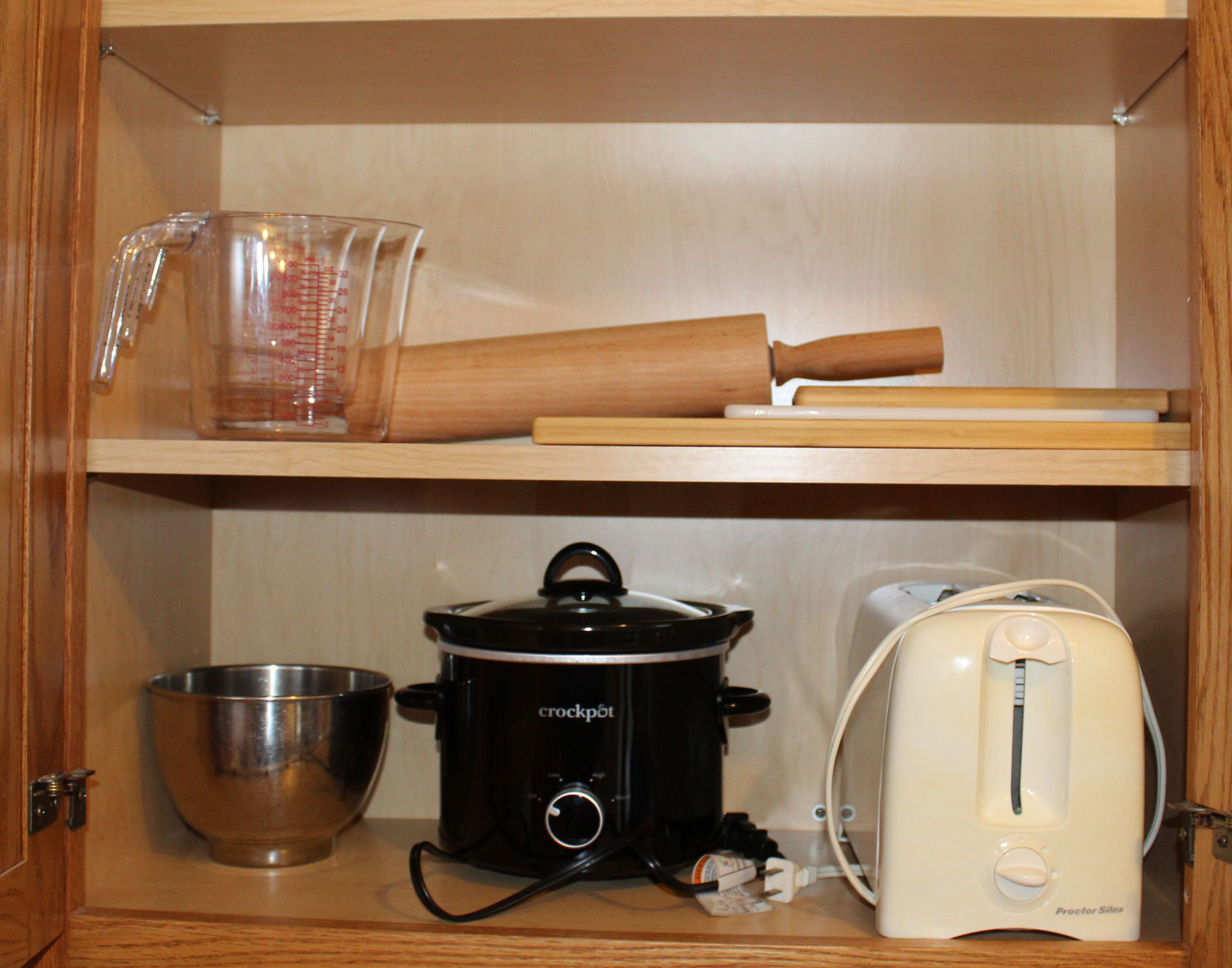 A kitchen with a stainless steel mixing bowl, a black crockpot, a white toaster, and a wooden rolling pin on a shelf.