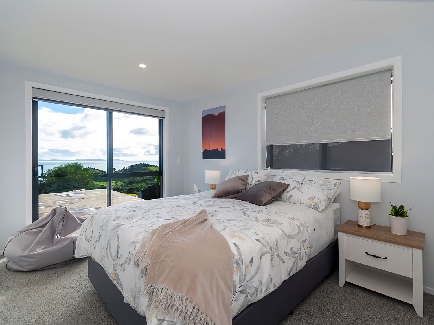 A modern bedroom with a large bed, a sliding glass door leading to a balcony, a bedside table with a lamp, and a wall-mounted TV.