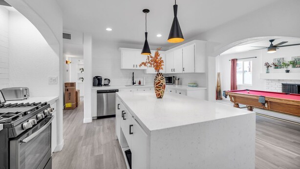 Una cocina moderna con armarios blancos, una isla central, lámparas colgantes negras y una mesa de billar.