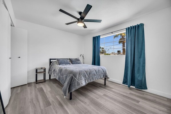A bedroom with a bed, a ceiling fan, a window with curtains, and a small table.