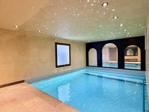 Piscina interior com água cristalina, paredes revestidas de azulejos e teto com iluminação embutida.
