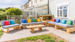 A patio area with wooden benches and cushions, a wooden table, and a glass-enclosed structure. - Coastguard Cottage (Atherfield)