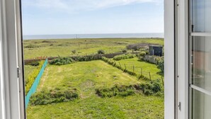A view from a room with a sliding glass door to a grassy outdoor area with a fence, a small shed, and a distant view of the sea. - Coastguard Cottage (Atherfield)