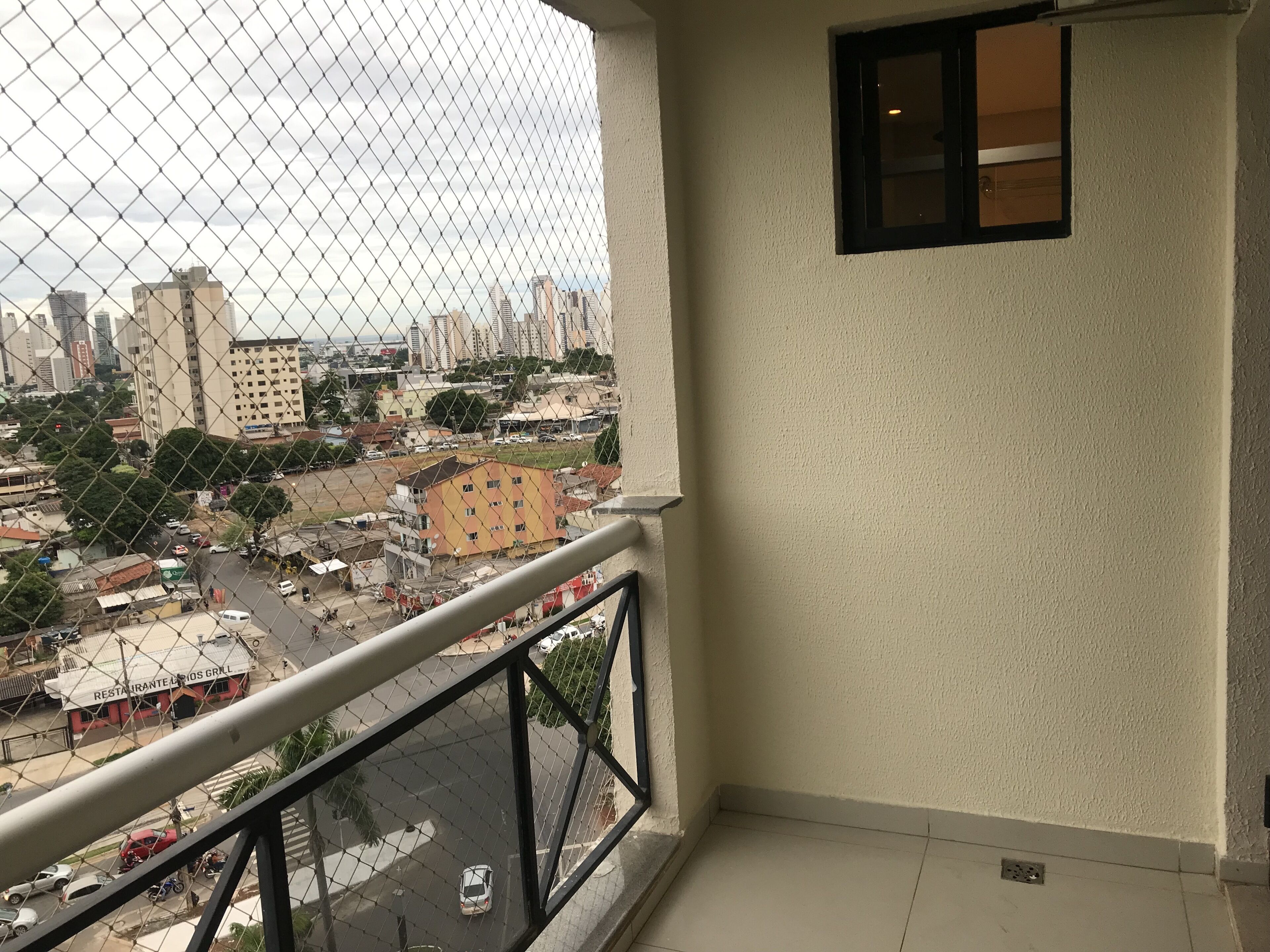 Un balcón con una cerca de malla metálica que da a una vista urbana, incluyendo edificios y una carretera.