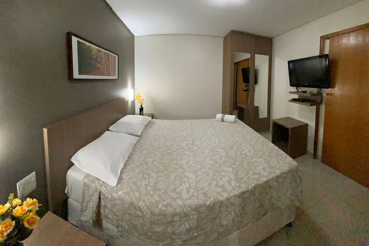 Une chambre d’hôtel avec un grand lit, une télévision, un miroir et une petite table agrémentée de fleurs.