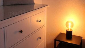 Ein Nachttisch mit Lampe, eine Schublade mit einem Kissen mit dem Aufdruck 'Lieblingsplatz' und eine weiße Kommode mit Schubladen.