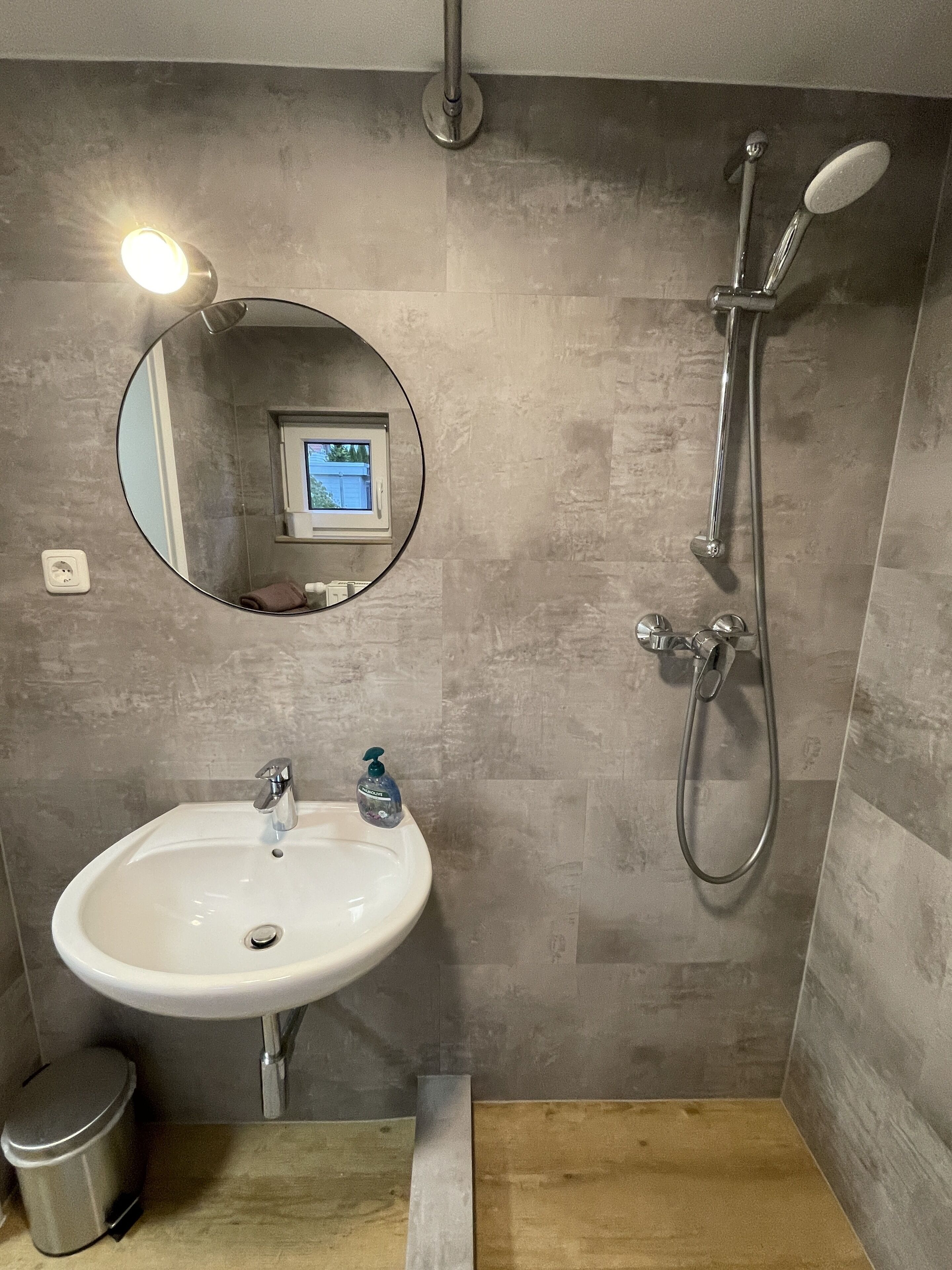 Une salle de bain avec un lavabo blanc, un miroir rond, une douchette à main et une fenêtre donnant sur un beau paysage.