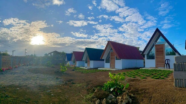 A row of modern, single-story cabins with pitched roofs, set on a dirt ground with sparse vegetation.