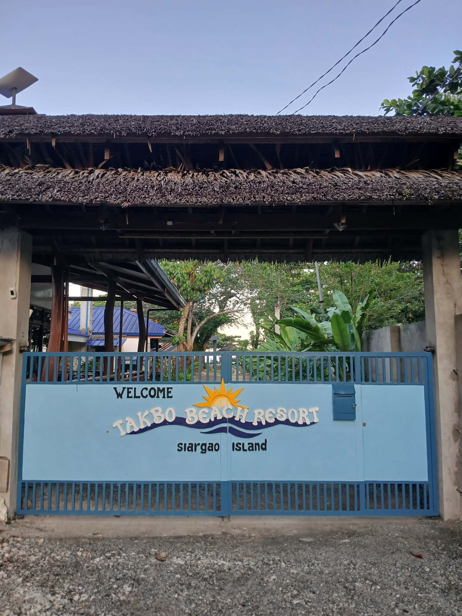 A blue gate with the text "Welcome Takibo Beach Resort Siargao Island" and a sun emblem.