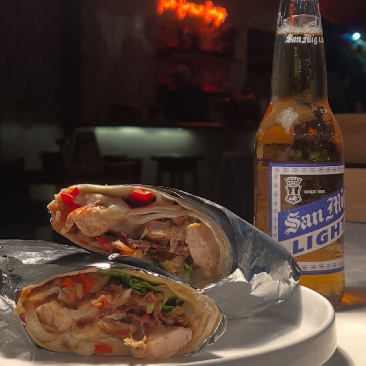 A burrito on a plate with a bottle of San Mig Light beer in the background.