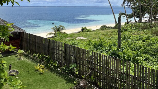 Vista di una spiaggia tropicale con una recinzione in legno, vegetazione rigogliosa e un cielo azzurro limpido.
