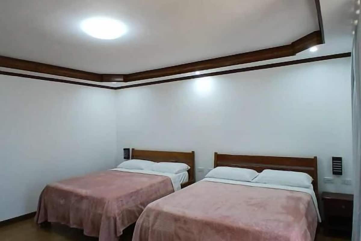 A small, clean room with two single beds, wooden flooring, white walls, and a simple ceiling design.