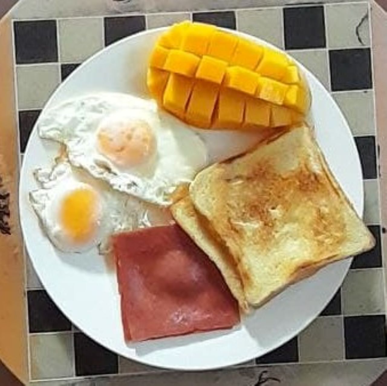 Een bord met twee gebakken eieren, een sneetje toast, een plakje ham en een halve mango.