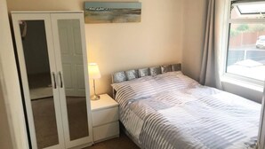 A bedroom with a bed, a nightstand, a wardrobe, a painting, and a window. - Lovely 3 Bedroom Holiday Home With Free Parking (Fareham)