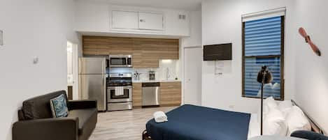 A compact room with a bed, a sofa, a small kitchen area, and a TV mounted on the wall.