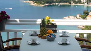 Ett bord dukat för fyra personer med vita koppar och fat, en ananas, en skål med frukt och blommor på en balkong med utsikt över ett kustnära område med byggnader och en strand.