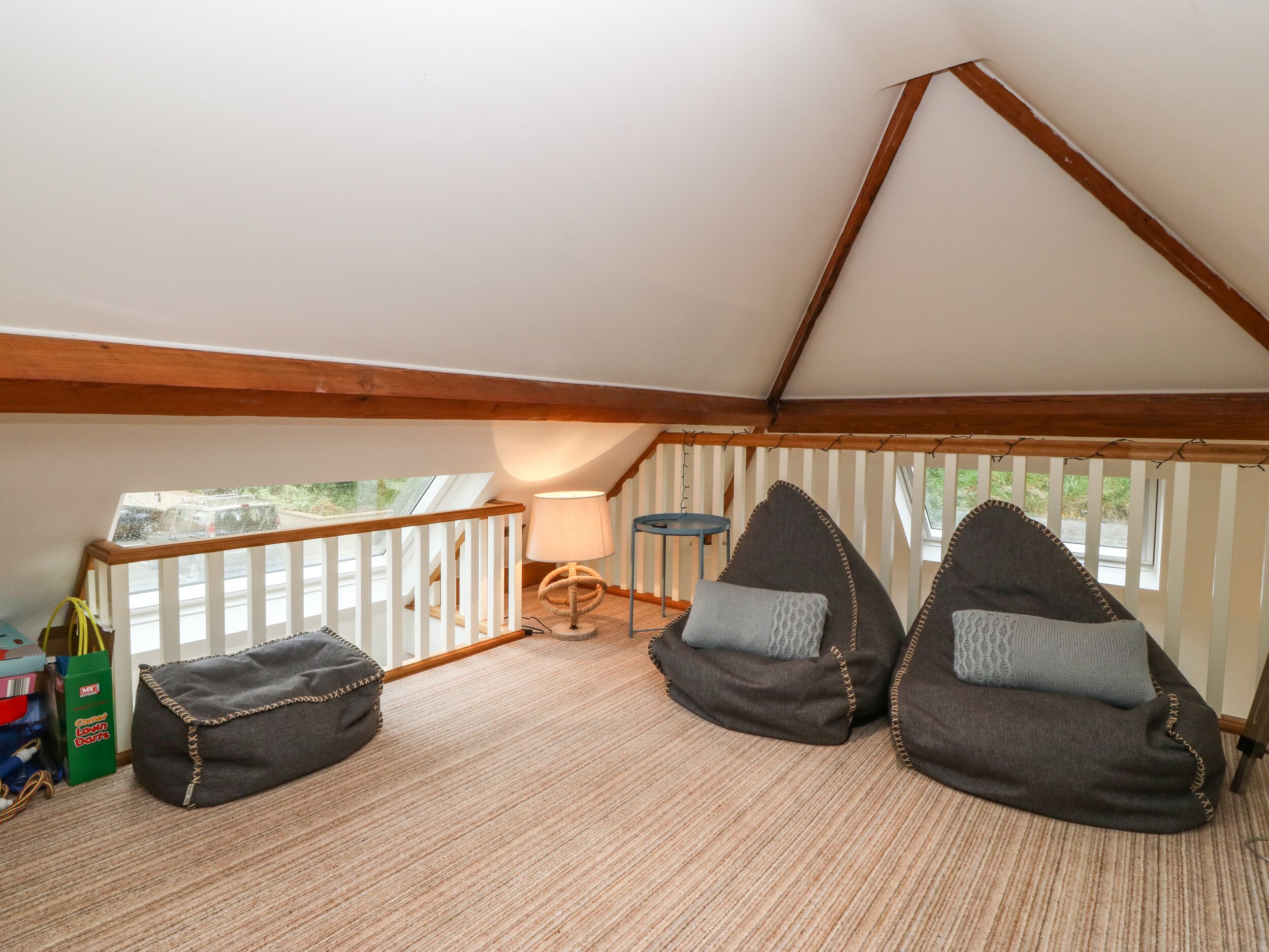 Una habitación acogedora en el desván, con techo inclinado, vigas de madera y vistas por una ventana. Hay dos pufs y una mesita con una lámpara.