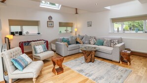 A spacious living room with a large grey sofa, a smaller armchair, and a coffee table. There are two windows providing natural light, and a wooden floor.