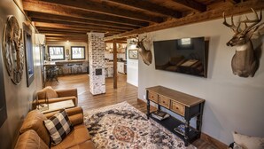 Un salon chaleureux avec un canapé en cuir marron, un tapis à motifs et une table basse en bois. Une grande tête de cerf est accrochée au mur, et on peut apercevoir un téléviseur.