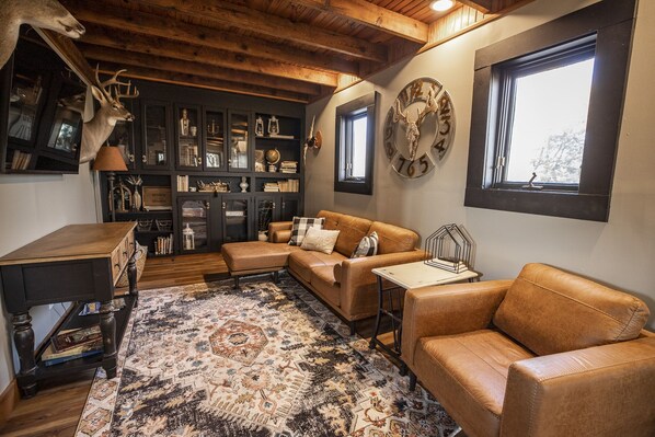 A cozy living room with a leather sofa, a coffee table, and a patterned rug. There is a wooden ceiling and a window with a view. - The Guesthouse Getaway (Lindsborg)