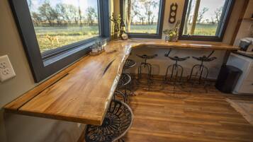A kitchen with a wooden countertop, metal bar stools, and a view of a grassy field through large windows.