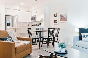 A modern living room with a sofa, a dining table with chairs, a kitchen area with a refrigerator and microwave, and a small coffee table with a remote control.