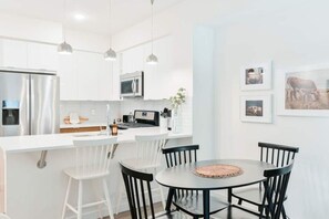 Eine moderne Küche mit weißen Schränken, einem Edelstahl-Kühlschrank, einer Mikrowelle und einem Essbereich mit rundem Tisch und schwarzen Stühlen.