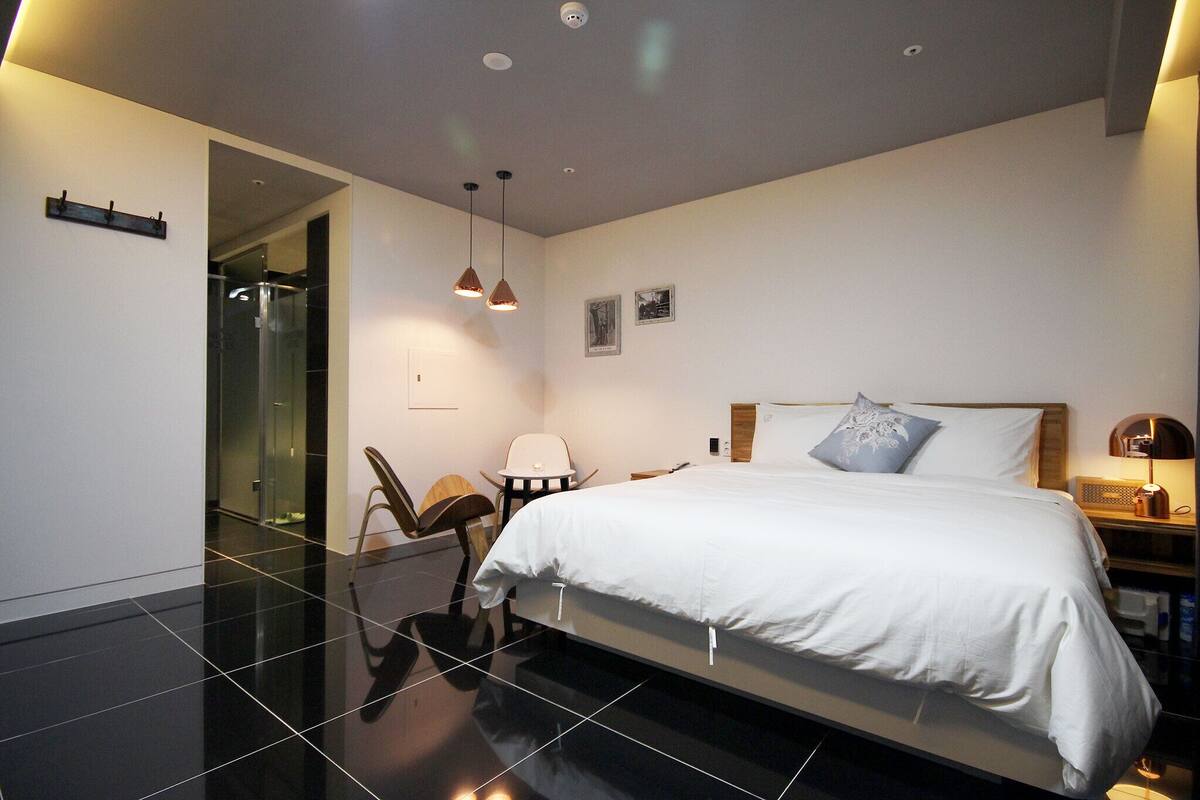 Un dormitorio moderno con una cama grande, una mesita de noche, una silla y un escritorio. Hay una puerta que da a un baño y una zona de ducha con mampara de cristal.
