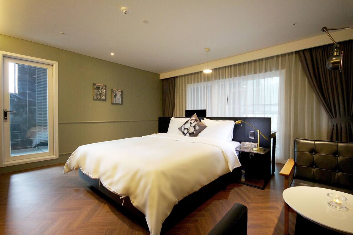 Un cuarto de hotel con una cama grande, una mesita de noche con lámpara, una silla y vistas al exterior a través de una ventana.