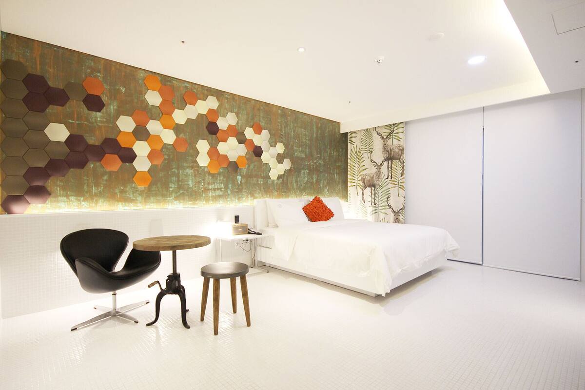 Un dormitorio moderno con una cama grande, una silla negra, una mesita redonda pequeña y una obra de arte hexagonal en la pared.