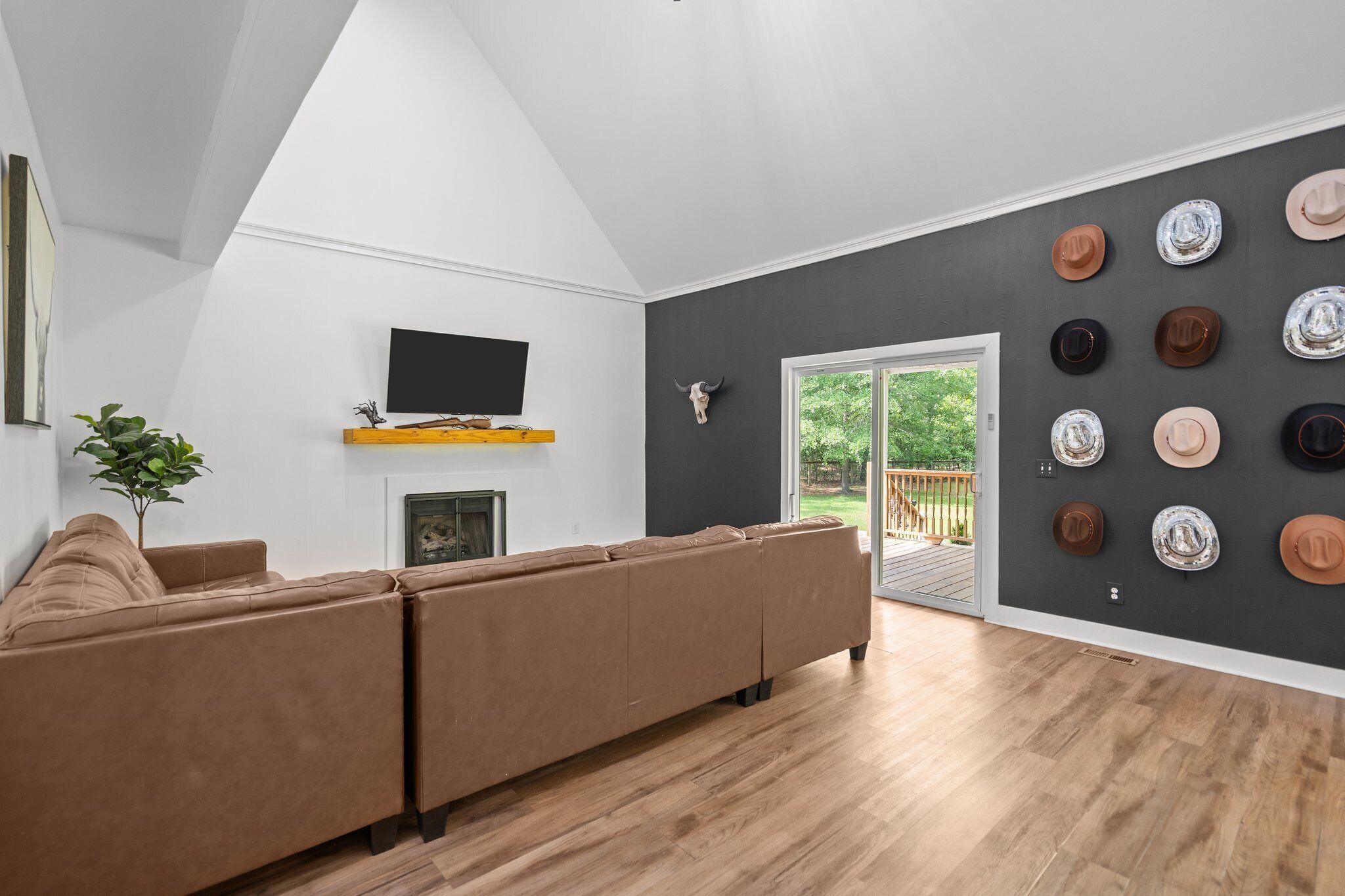 Una sala de estar moderna con un sofá seccional marrón, un televisor de pantalla plana sobre un estante de madera, una decoración de cráneo montado y una pared con varios sombreros.
