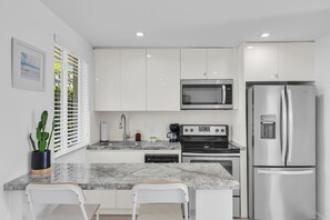 A modern kitchen with white cabinets, a stainless steel refrigerator, a built-in microwave, and a stove. There is a marble countertop and two white chairs.