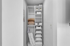 A white closet with shelves for folded clothes, a hanging rack for towels, and a shelf with rolled towels and potted plants.
