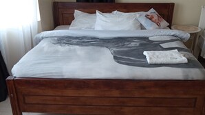 A wooden bed with a grey and white patterned bedspread, white pillows, and a folded white towel on the bed.