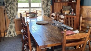 Taula de dinar de fusta amb caires, un ordinador portàtil i un quadern. Hi ha una finestra amb cortines, un armari de fusta amb vidrieria, i una paret de pedra al fons.