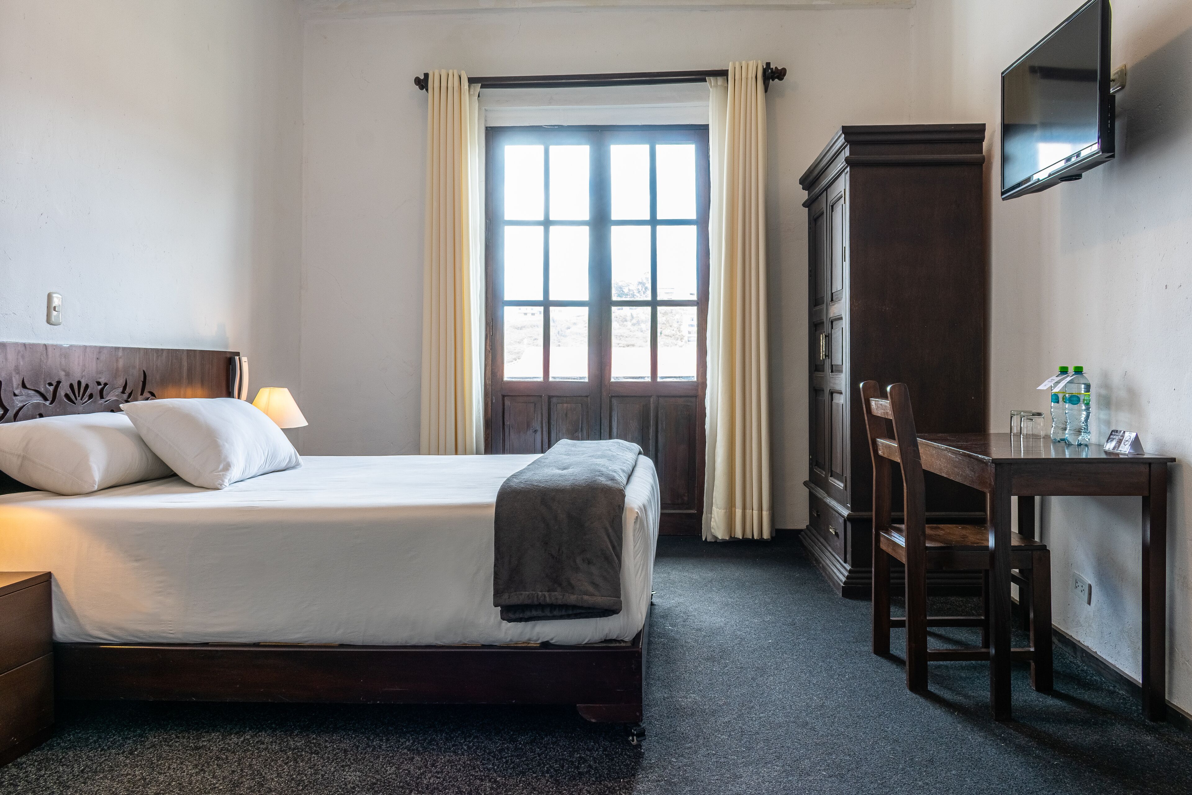 Habitación de hotel con cama, escritorio, silla, armario y ventana con cortinas.