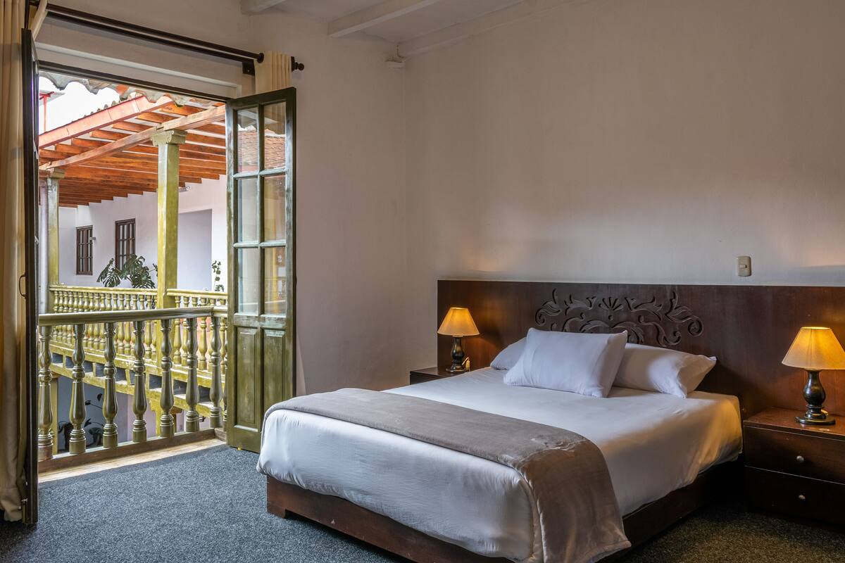 Une chambre d’hôtel avec un grand lit, deux lampes de chevet, une tête de lit en bois et un balcon avec une balustrade.