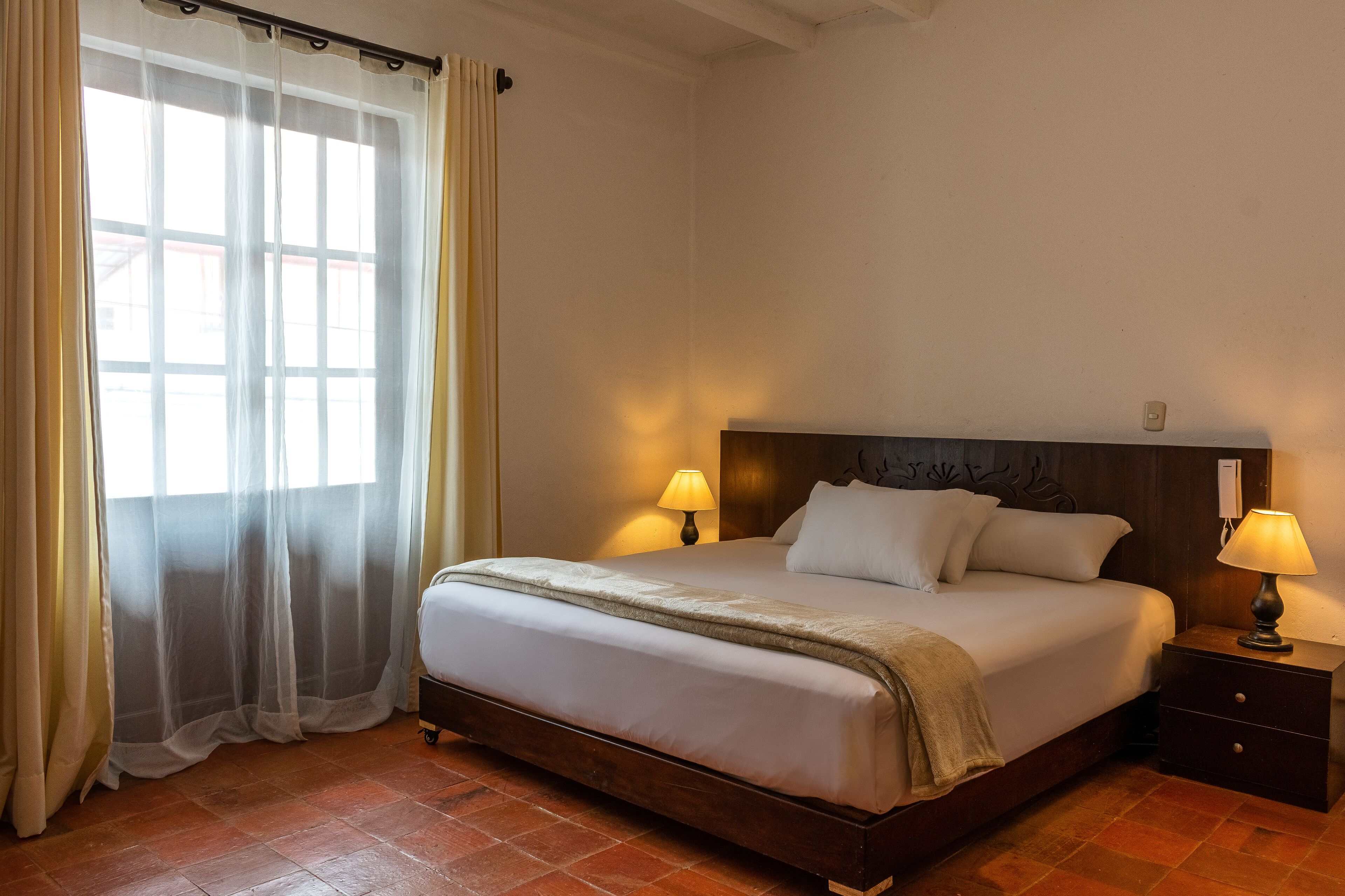 A neatly made bed with a wooden headboard, two bedside lamps, and a window with sheer curtains.