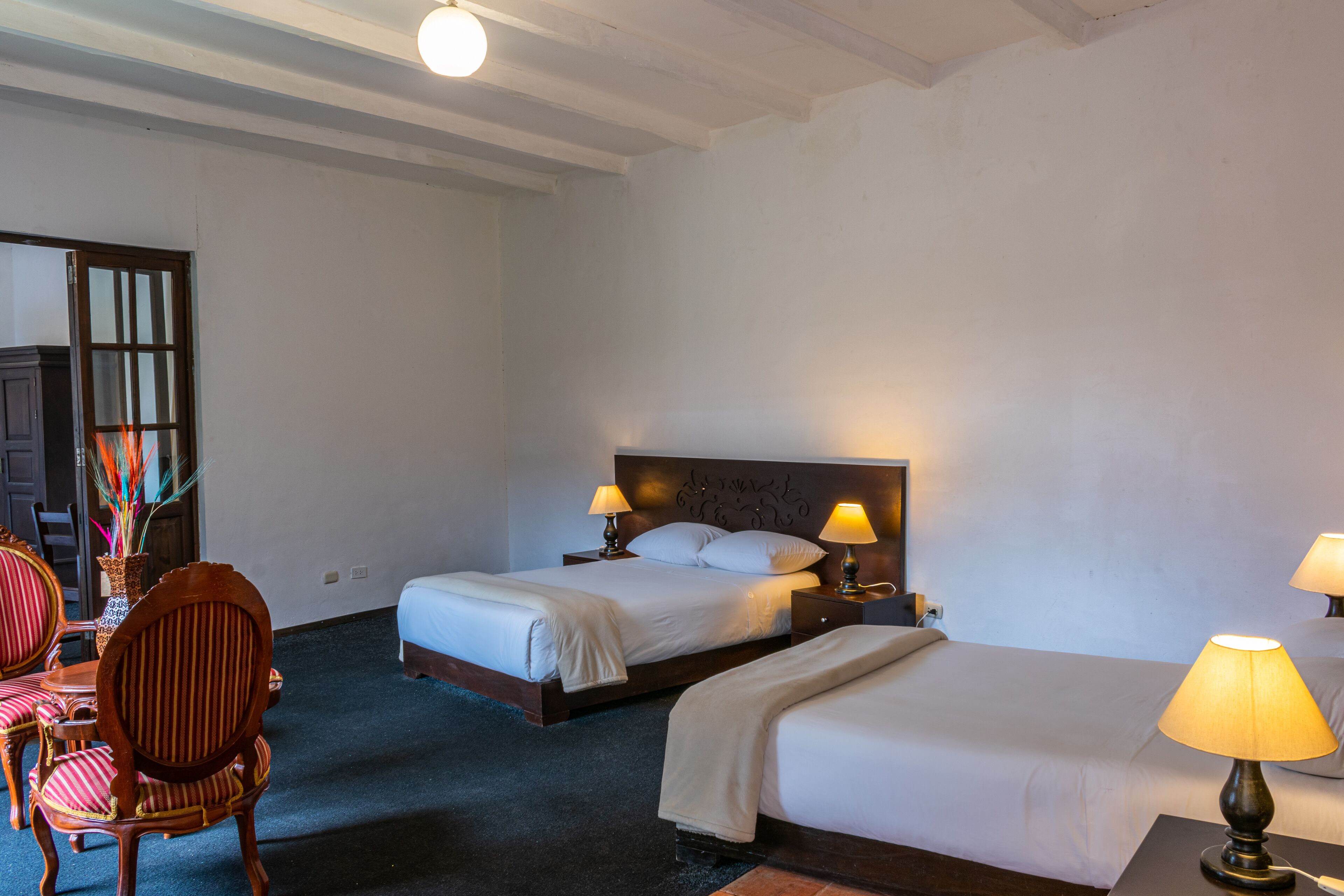 Une chambre d’hôtel avec deux lits, une tête de lit en bois, une table de chevet avec des lampes, une chaise et un vase avec des fleurs.