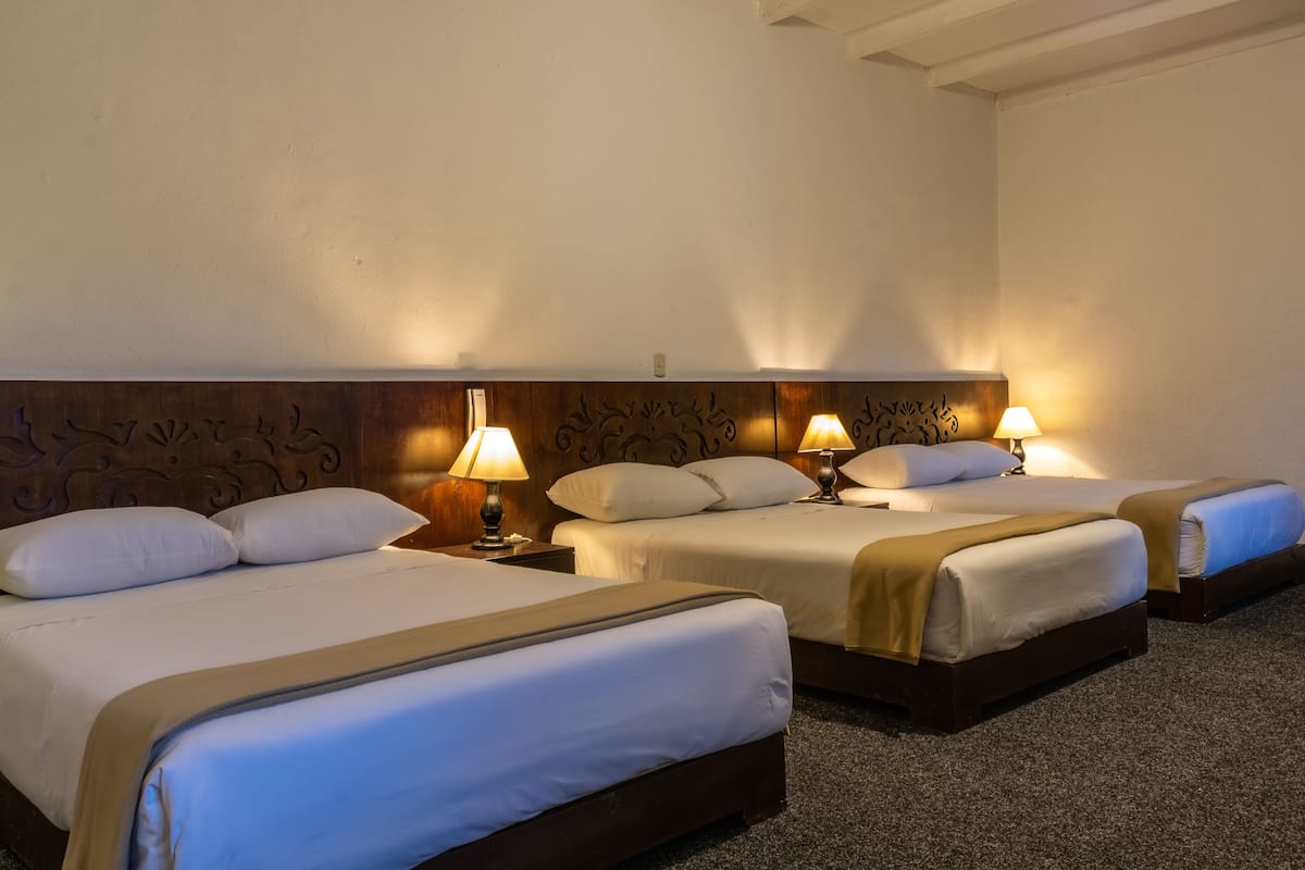 Une chambre d’hôtel avec trois lits, chacun doté de draps blancs et d’une tête de lit marron. Chaque lit est muni d’une lampe de chevet.