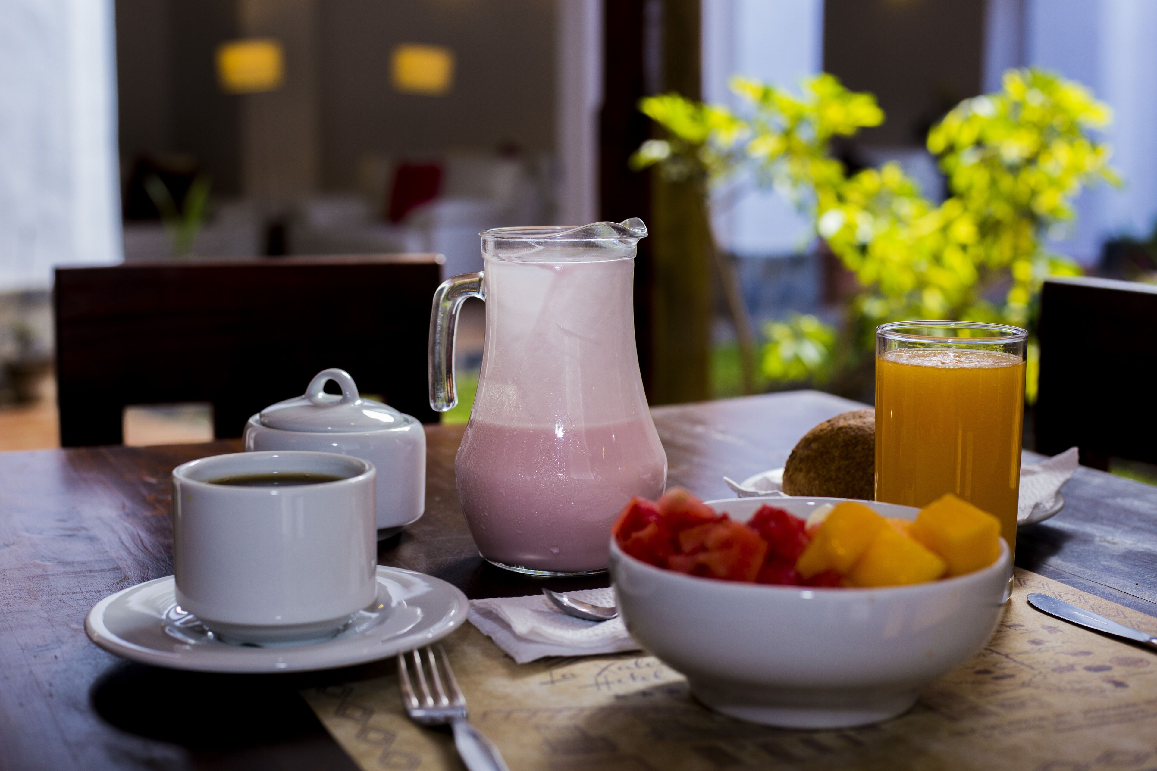 A breakfast setting with a pitcher of pink smoothie, a glass of orange juice, a bowl of fruit, and a cup of coffee on a wooden table.